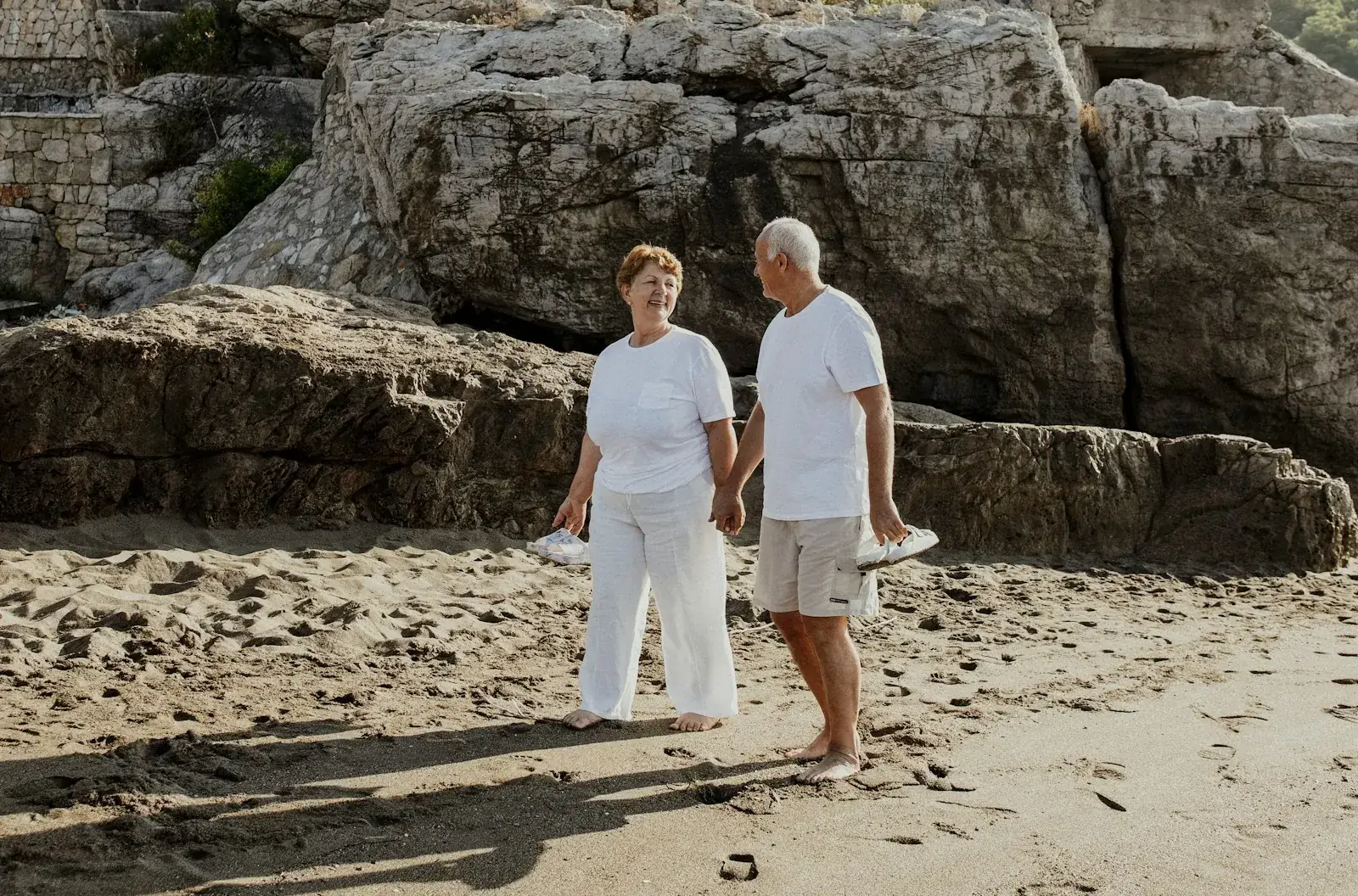 An older couple walking hand in hand along a sandy beach beneath rocky cliffs.