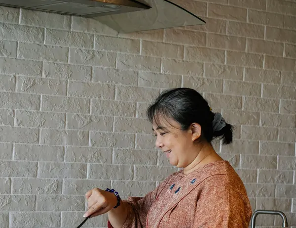 An older woman smiling as she cooks at the stove in a bright home kitchen.