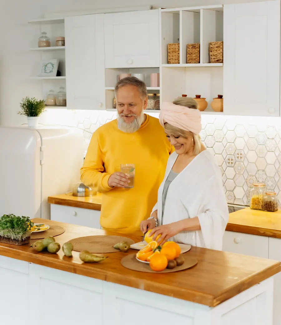 An older couple laughing together while preparing fresh fruit in a bright kitchen.