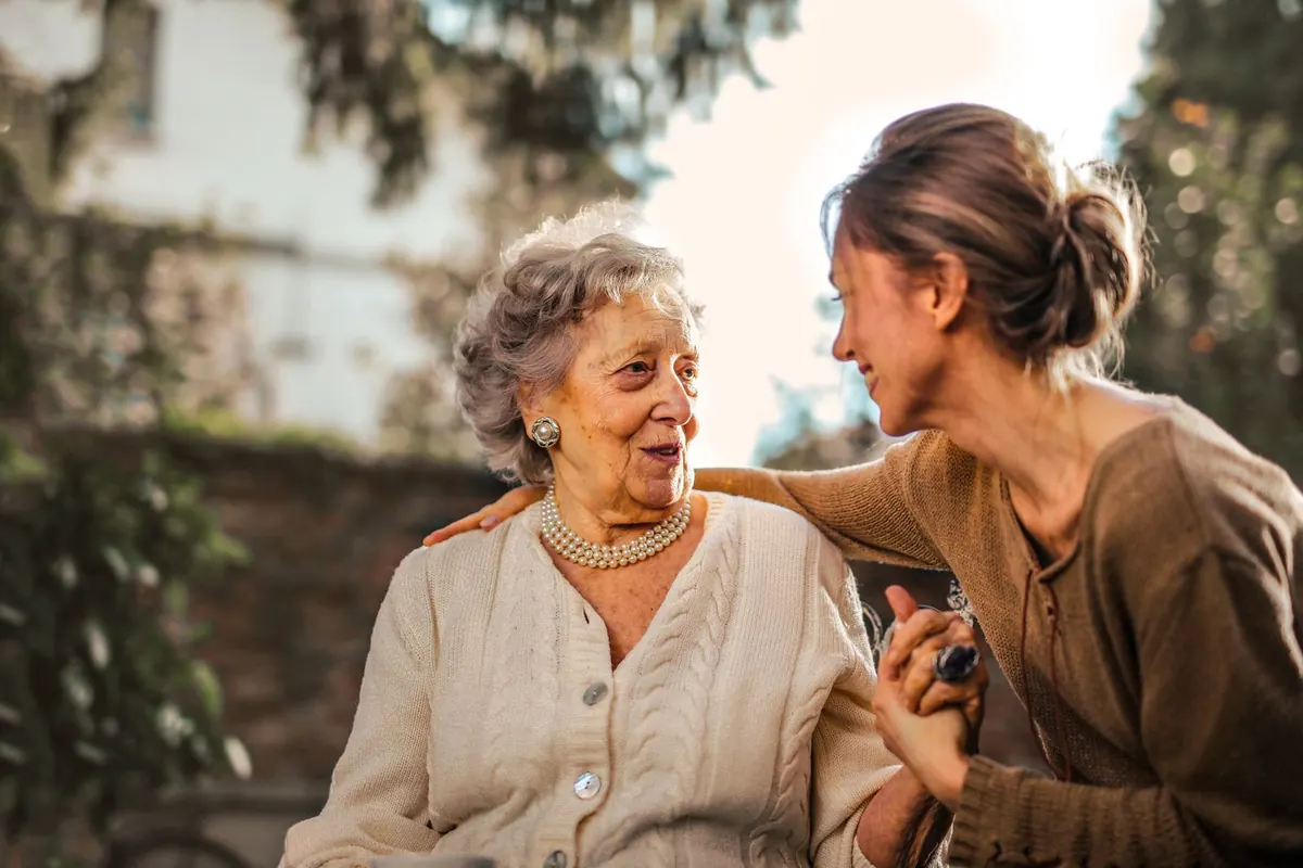 An older woman and her adult daughter laughing together in a sunny garden.