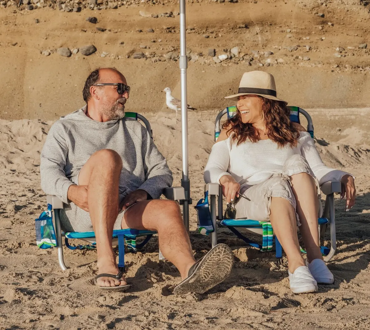 An older couple in sun hats relaxing in beach chairs on the sand, chatting and smiling.