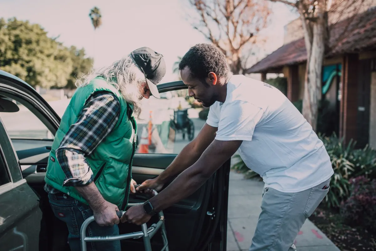 A younger man helping an older man with a walking frame as he steps out of a car on a sunny street.